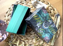 Load image into Gallery viewer, Meraki tarot 3rd edition open box. Cards in the box are on their side to show green edging. Everything is laying on a wicker mat with crystals on a wood background