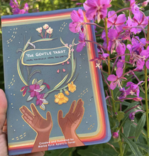 Load image into Gallery viewer, Hand holding the Gentle Tarot guidebook in front of purple flowers and greenery