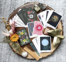 Load image into Gallery viewer, The cards of the Terra Lenormand deck in a decorative arrangement with flowers and crystals set atop a cross section of a tree trunk