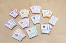 Load image into Gallery viewer, Sets of cards from the Vulva Quartet game sorted by color and lying on a wood background