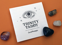 Load image into Gallery viewer, Front of Trinity Tarot prompt sheet laying on an orange background with 4 stones surrounding it