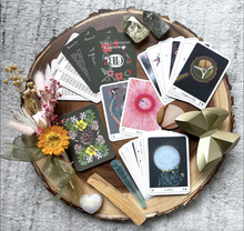 Load image into Gallery viewer, The cards of the Terra Lenormand deck in a decorative arrangement with flowers and crystals set atop a cross section of a tree trunk
