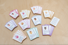 Load image into Gallery viewer, Sets of cards from the Vulva Quartet game sorted by color and lying on a wood background
