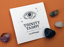 Load image into Gallery viewer, Front of Trinity Tarot prompt sheet laying on an orange background with 4 stones surrounding it
