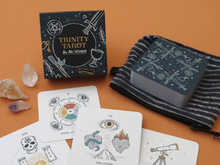 Load image into Gallery viewer, Display on an orange background consisting of the Trinity Tarot tuck box, 4 face up cards, the rest of the deck stacked and face down on a black and gray striped cloth and 3 crystals along the left side
